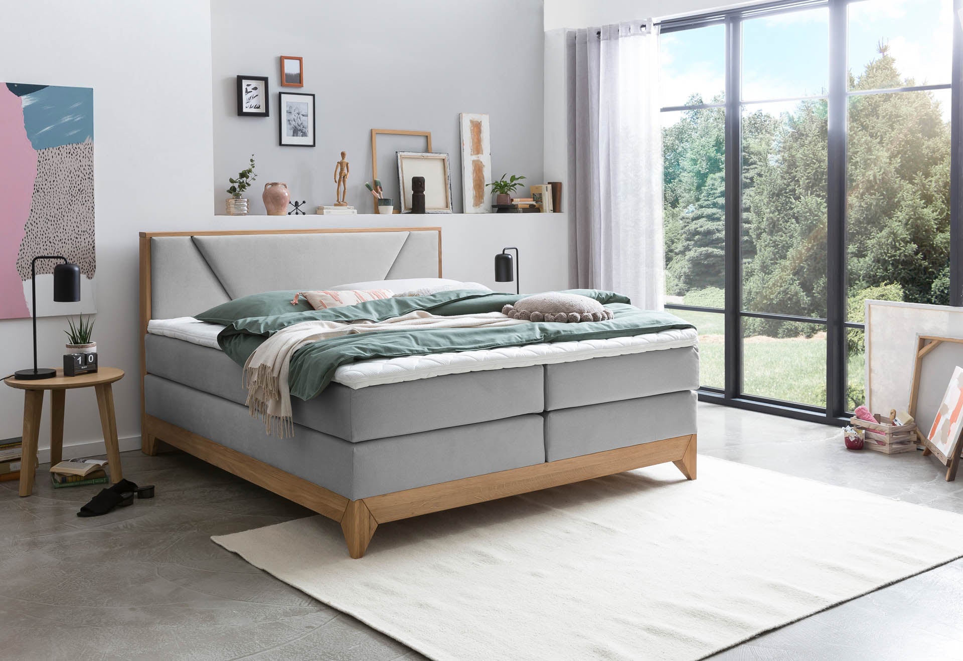 Helles Schlafzimmer mit grauem Boxspringbett auf Holzrahmen, Topper und Bettwäsche sowie dekorative Elemente wie Bildern, Pflanzen und Wohnaccessoires