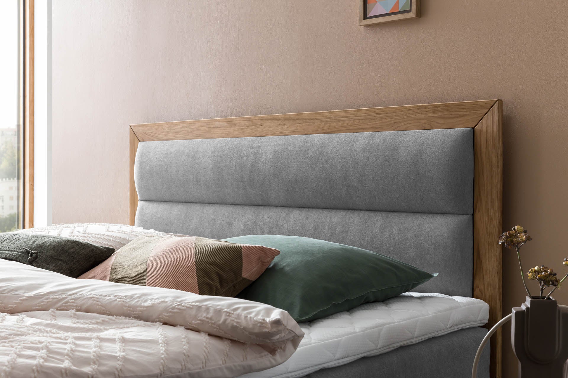Boxspringbett mit grauem, gepolstertem Kopfteil und markantem Holzrahmen in modernem Schlafzimmer mit warmen Wandfarben. 